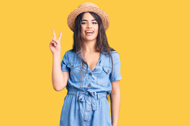 Brünettes Teenager-Mädchen mit Sommermütze zeigt und zeigt mit Fingern Nummer zwei nach oben, während sie selbstbewusst und glücklich lächelt.  - Foto, Bild