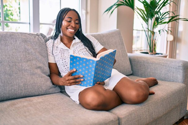 Joven afroamericana americana sonriendo feliz lectura libro en casa - Foto, Imagen