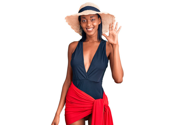 Jovem afro-americana vestindo maiô e chapéu de verão mostrando e apontando para cima com os dedos número cinco enquanto sorri confiante e feliz.  - Foto, Imagem