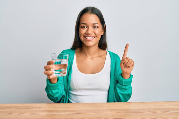 Beautiful hispanic woman drinking glass of water smiling with an idea or question pointing finger with happy face, number one  - Fotografie, Obrázek