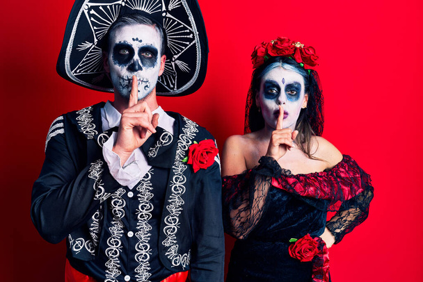 Young couple wearing mexican day of the dead costume over red asking to be quiet with finger on lips. silence and secret concept.  - Φωτογραφία, εικόνα