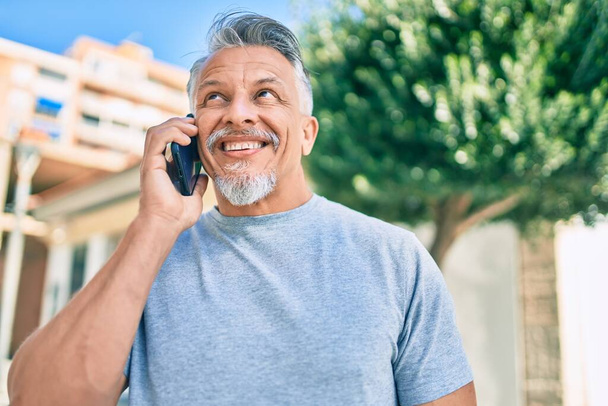 Латиноамериканец средних лет седой мужчина улыбается счастливо разговаривая по смартфону в городе. - Фото, изображение