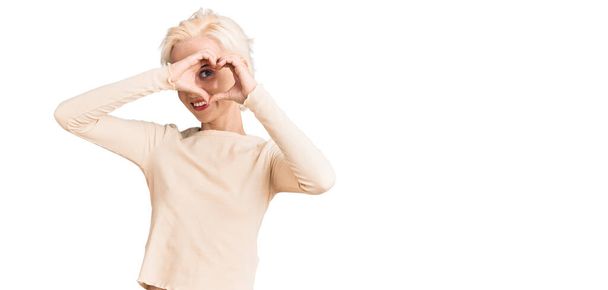 Молодая блондинка в повседневной одежде делает форму сердца с рукой и пальцами, улыбаясь, глядя через знак  - Фото, изображение