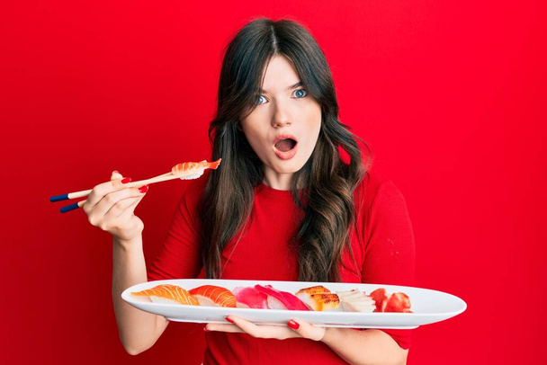 Nuori kaunis valkoihoinen tyttö syö sushia käyttäen syömäpuikkoja shokki kasvot, etsivät skeptinen ja sarkastinen, yllättynyt suu auki  - Valokuva, kuva