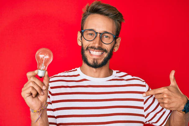 Schöner kaukasischer Mann mit Bart, der eine Glühbirne als Inspiration und Idee hält und mit dem Finger auf einen selbst lächelnd glücklich und stolz zeigt  - Foto, Bild