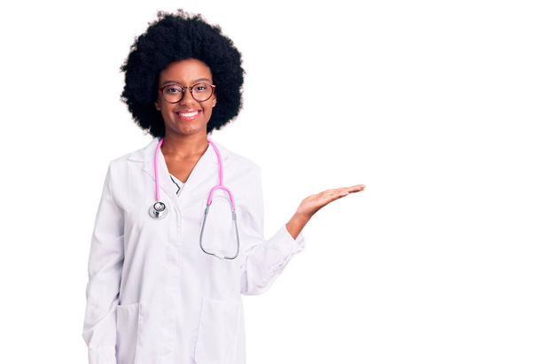 Fiatal afro-amerikai nő orvos kabát és sztetoszkóp mosolygós vidám bemutató és mutató tenyérrel a kamera.  - Fotó, kép
