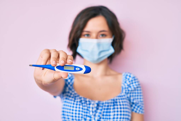 Young beautiful woman wearing medical mask holding holding thermometer thinking attitude and sober expression looking self confident  - Photo, Image