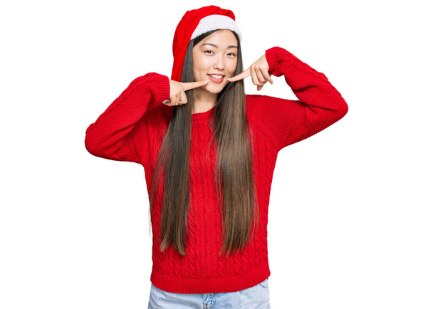 Mladá Číňanka s vánočním kloboukem vesele se usmívající a ukazující prstem na zuby a ústa. koncepce stomatologického zdraví.  - Fotografie, Obrázek