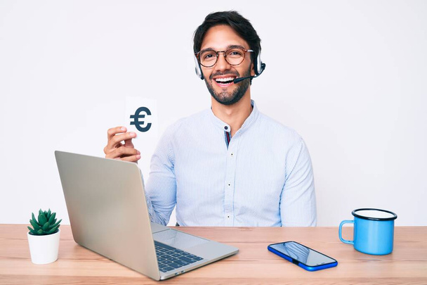 Handsome hispanic man working at the office holding euro banner looking positive and happy standing and smiling with a confident smile showing teeth  - Foto, imagen