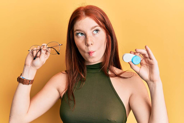 Young red head girl holding glasses and contact lenses making fish face with mouth and squinting eyes, crazy and comical.  - Photo, Image