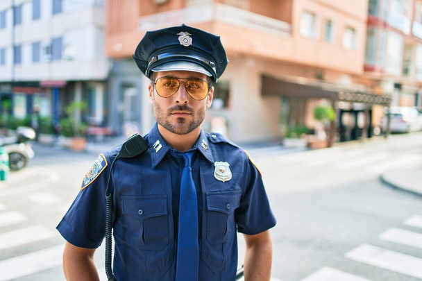 Young handsome hispanic policeman wearing police uniform. Standing with serious expression at town street. - Photo, Image