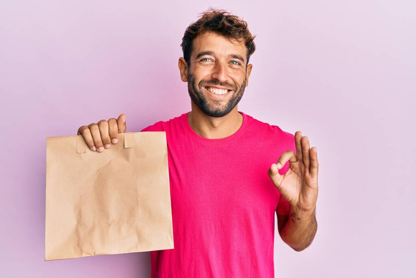 Handsome man with beard holding take away paper bag doing ok sign with fingers, smiling friendly gesturing excellent symbol  - Foto, Bild