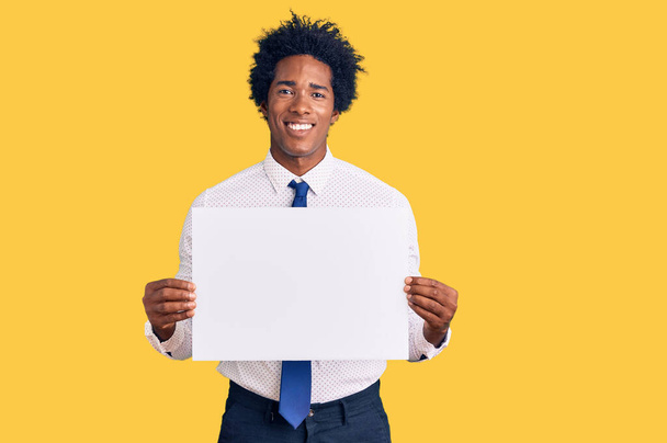 Handsome african american man with afro hair holding blank empty banner looking positive and happy standing and smiling with a confident smile showing teeth  - 写真・画像