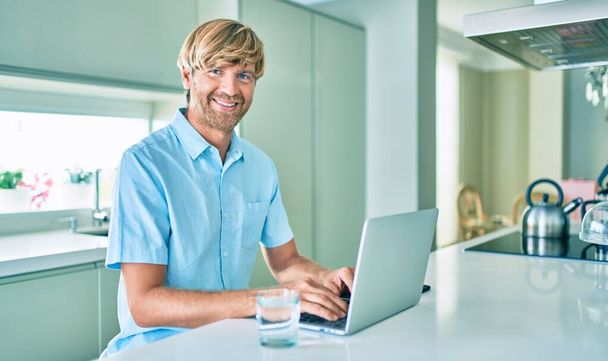 Молодой ирландец улыбается счастливой работе с ноутбуком, сидя дома на столе. - Фото, изображение