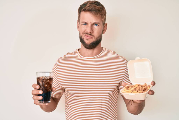 Young caucasian man eating fast food clueless and confused expression. doubt concept.  - Φωτογραφία, εικόνα