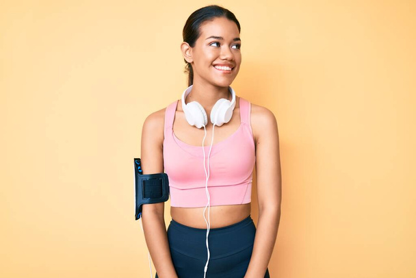 Young beautiful latin girl wearing gym clothes and using headphones looking to side, relax profile pose with natural face and confident smile.  - Zdjęcie, obraz