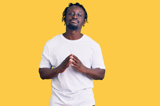 Mladý afro-americký muž s copánky na sobě ležérně bílé tričko ruce k sobě a prsty zkřížené úsměv uvolněně a vesele. úspěch a optimismus  - Fotografie, Obrázek