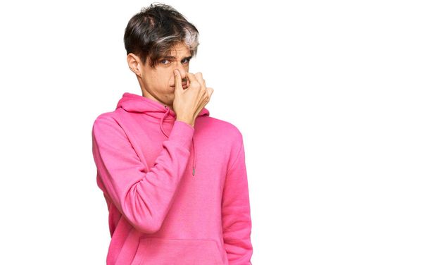 junger hispanischer Mann in lässigem rosafarbenem Sweatshirt, der etwas Stinkendes und ekelhaften, unerträglichen Geruch riecht und mit den Fingern auf der Nase den Atem anhält. schlechter Geruch  - Foto, Bild