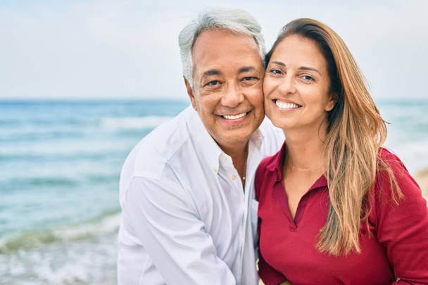 Latynoska para w średnim wieku uśmiecha się szczęśliwa i przytula spacerując po plaży - Zdjęcie, obraz
