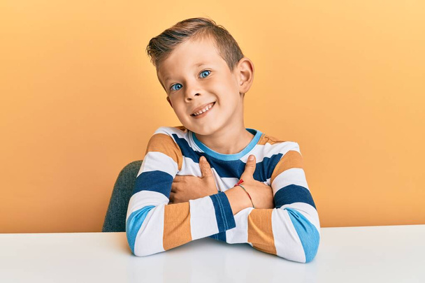 Αξιολάτρευτο καυκάσιο παιδί που φοράει καθημερινά ρούχα κάθεται στο τραπέζι χαρούμενο πρόσωπο χαμογελώντας με σταυρωμένα χέρια κοιτάζοντας την κάμερα. θετικό πρόσωπο.  - Φωτογραφία, εικόνα