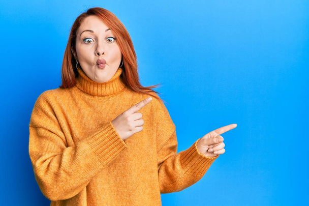 Beautiful redhead woman pointing fingers to the side making fish face with mouth and squinting eyes, crazy and comical.  - Fotoğraf, Görsel