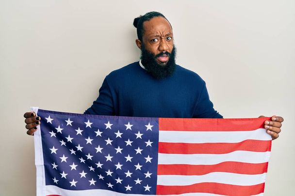 Junger afrikanisch-amerikanischer Mann mit einer Flagge der Vereinigten Staaten, ahnungslos und verwirrt. Zweifelhaftes Konzept.  - Foto, Bild