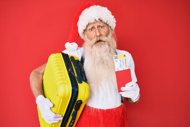 Old senior man wearing santa claus costume holding suicase and boarding pass in shock face, looking skeptical and sarcastic, surprised with open mouth  - 写真・画像