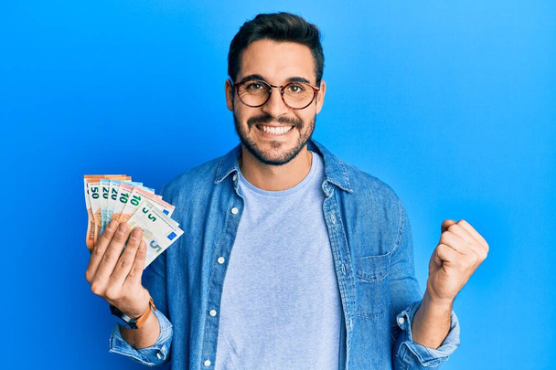 Junger hispanischer Mann hält stolz Euro-Banknoten in der Hand und feiert Sieg und Erfolg aufgeregt mit erhobenem Arm  - Foto, Bild