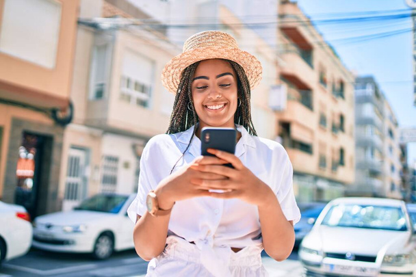 Jeune femme afro-américaine avec des tresses souriant heureux en utilisant smartphone à l'extérieur par une journée ensoleillée de l'été - Photo, image
