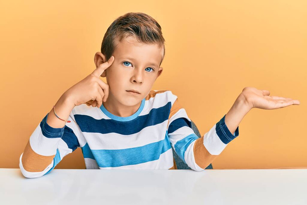 Adorable caucasian kid wearing casual clothes sitting on the table confused and annoyed with open palm showing copy space and pointing finger to forehead. think about it.  - Photo, Image