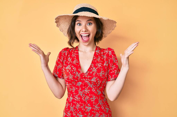 Mladá krásná dívka v letním klobouku slaví vítězství se šťastným úsměvem a vítězným výrazem se zdviženýma rukama  - Fotografie, Obrázek