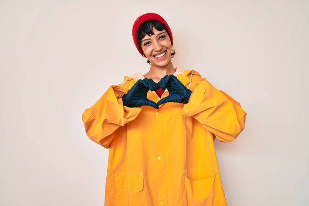 Beautiful brunettte woman wearing yellow raincoat smiling in love showing heart symbol and shape with hands. romantic concept.  - Photo, Image