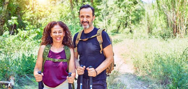 Beautiful couple of hiker wearing backpack smiling happy. Standing with smile on face doing trekking using hiking stick at forest - Fotó, kép