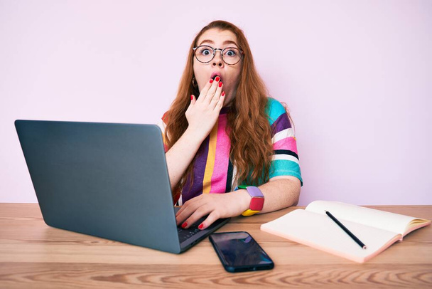 Jonge roodharige vrouw die op kantoor werkt met een laptop die mond met de hand bedekt, geschokt en bang voor fouten. verraste uitdrukking  - Foto, afbeelding