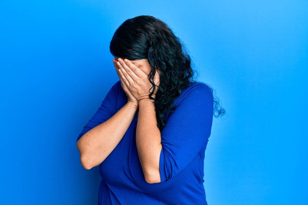 Συν μέγεθος μελαχρινή γυναίκα φορώντας casual μπλε πουκάμισο με λυπημένη έκφραση που καλύπτει το πρόσωπο με τα χέρια, ενώ κλαίει. έννοια της κατάθλιψης.  - Φωτογραφία, εικόνα