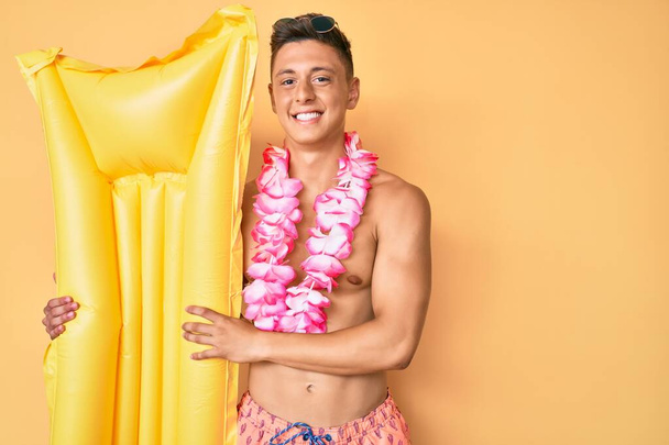Joven niño hispano usando traje de baño y sosteniendo colchón de verano flotan buscando positivo y feliz de pie y sonriendo con una sonrisa confiada mostrando los dientes  - Foto, Imagen