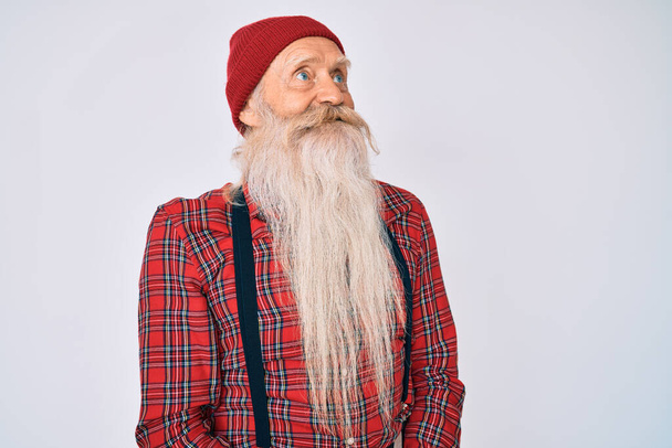 Old senior man with grey hair and long beard wearing hipster look with wool cap looking away to side with smile on face, natural expression. laughing confident.  - Foto, Imagem
