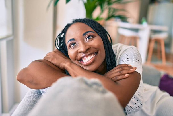 Νεαρή Αφρικάνα Αμερικανίδα χαμογελαστή χαρούμενη κάθεται στον καναπέ στο σπίτι - Φωτογραφία, εικόνα