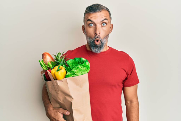 Middle age handsome man holding paper bag with bread and groceries scared and amazed with open mouth for surprise, disbelief face  - 写真・画像
