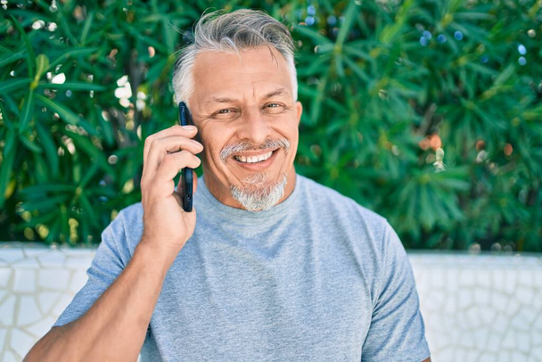 Латиноамериканец средних лет седой мужчина улыбается счастливо разговаривая по смартфону в парке. - Фото, изображение