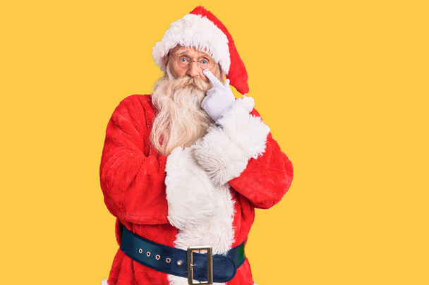 старик с седыми волосами и длинной бородой в традиционном костюме Санта-Клауса, указывая на глаз, наблюдая за твоим жестом, подозрительным выражением лица  - Фото, изображение