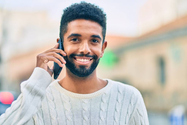 Joven hombre afroamericano sonriendo feliz hablando en el teléfono inteligente en la ciudad. - Foto, Imagen