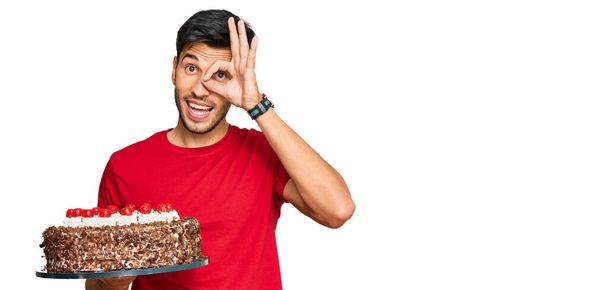 Молодой красивый мужчина празднует день рождения с тортом улыбается счастливо делает хорошо знак с рукой на глазу глядя сквозь пальцы  - Фото, изображение