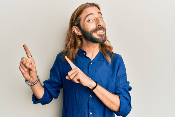Aantrekkelijke man met lang haar en baard in casual kleding glimlachend en kijkend naar de camera wijzend met twee handen en vingers naar de zijkant.  - Foto, afbeelding