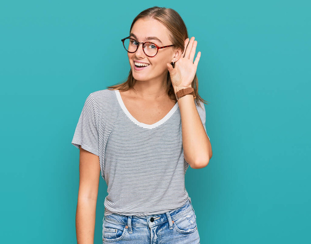 Belle jeune femme blonde portant des vêtements décontractés et des lunettes souriant main sur oreille en écoutant une rumeur ou des commérages. concept de surdité.  - Photo, image