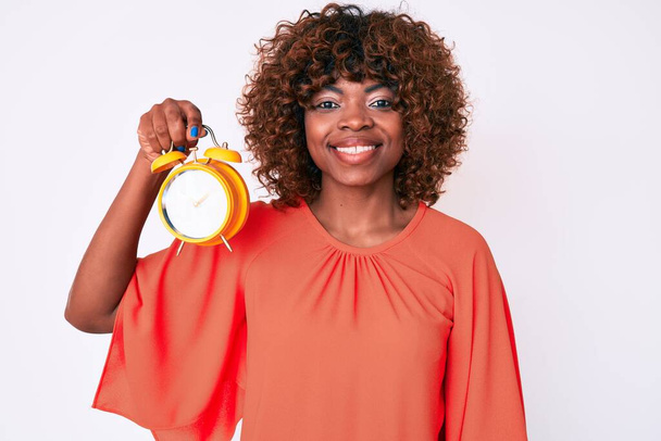 Young african american woman holding alarm clock looking positive and happy standing and smiling with a confident smile showing teeth  - Photo, Image