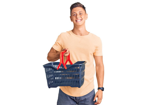 Jonge knappe Latijns-Amerikaanse man met supermarkt winkelmand op zoek positief en gelukkig staan en glimlachen met een zelfverzekerde glimlach tonen tanden  - Foto, afbeelding