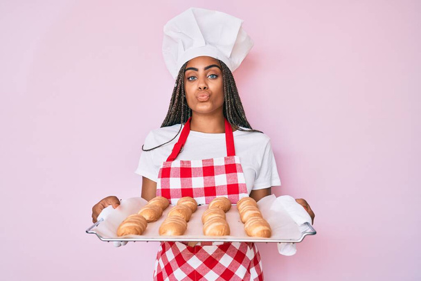Молодая африканская американка с косичками в форме пекаря, держащая домашний хлеб, смотрящая на камеру, дующую в поцелуй, будучи милой и сексуальной. выражение любви.  - Фото, изображение