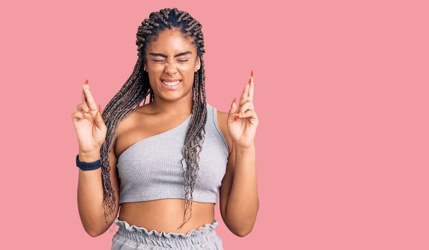 Молода афро-американська жінка з плечима, одягнена в спортивну лайку, схрестила палець, посміхаючись із заплющеними очима. удача і забобонне поняття.  - Фото, зображення