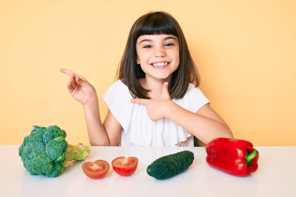 Kleines Mädchen mit Knall sitzt auf dem Tisch mit Gemüse lächelnd und blickt in die Kamera, die mit zwei Händen und Fingern zur Seite zeigt.  - Foto, Bild